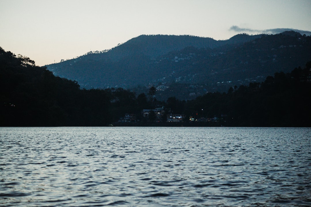 View of the Bhimtal lake at the time of sunset, also visible are the few cafeterias near the coast as well as small villages on the mountains. by Boat Brands a body of water with a hill in the background